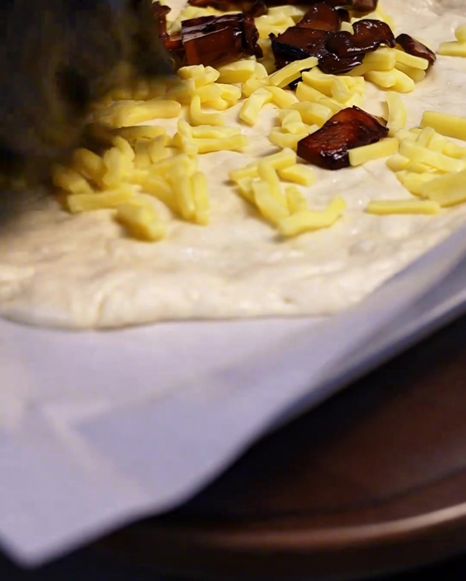 Cooked, seasoned mushrooms being spooned onto a layer of shredded cheese over a pizza dough base.