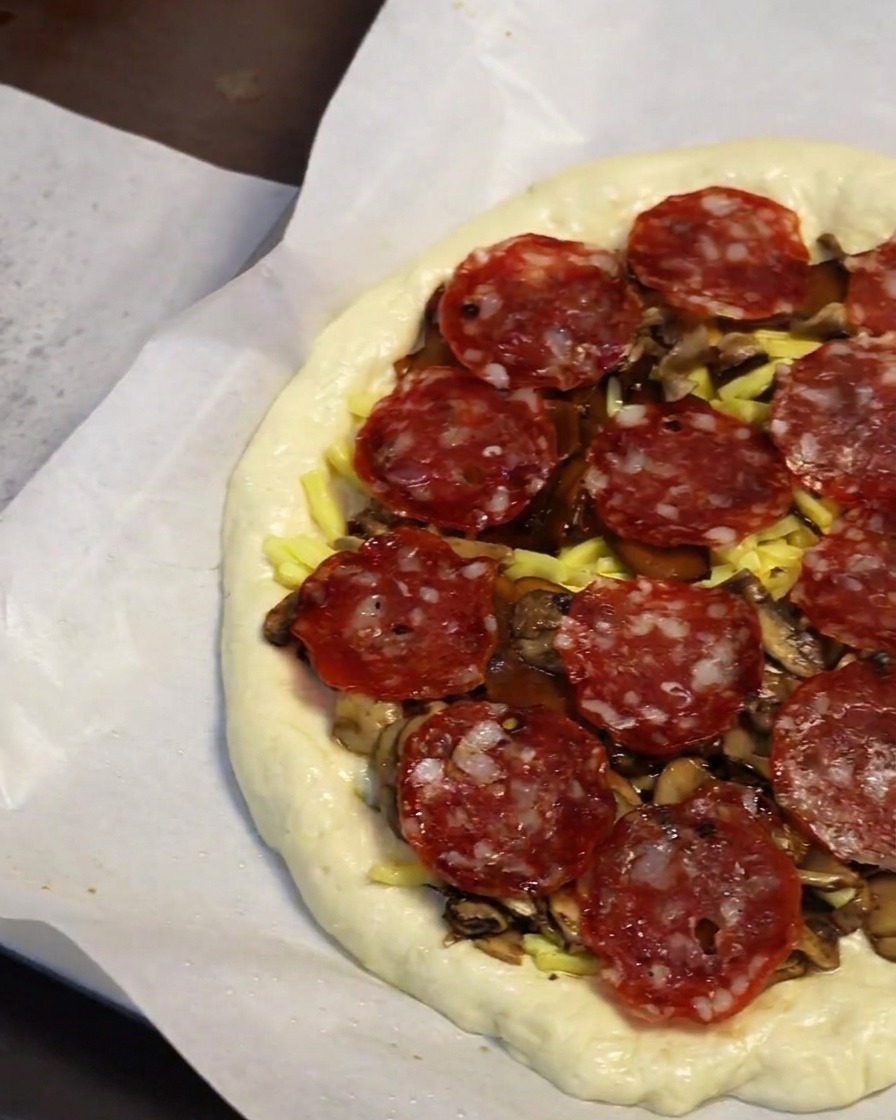 Close-up of a hand placing round pepperoni slices onto a layer of mushrooms and cheese on pizza dough.