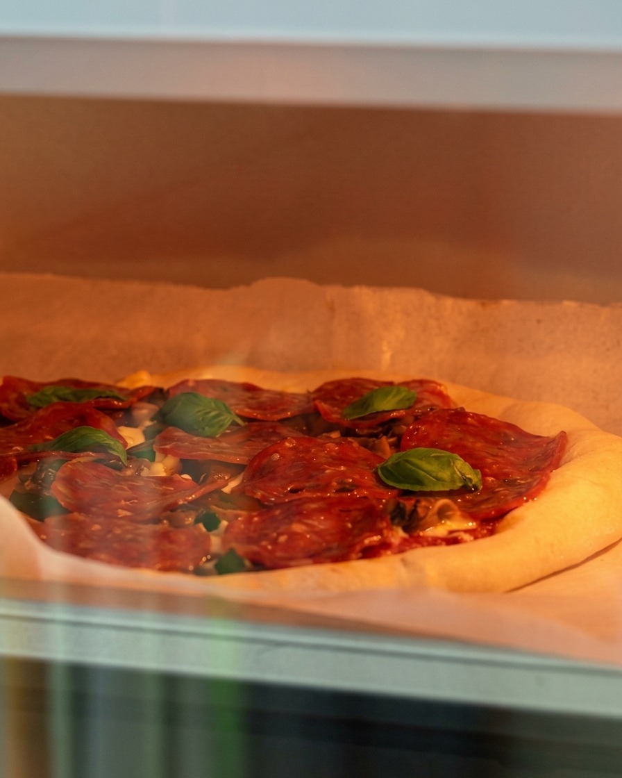 A raw pizza topped with cheese, mushrooms, and pepperoni being carefully placed into a hot oven.