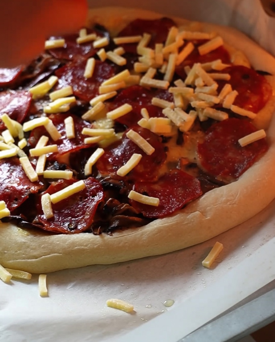 A partially baked pizza inside an oven receiving a generous final dusting of shredded cheese.