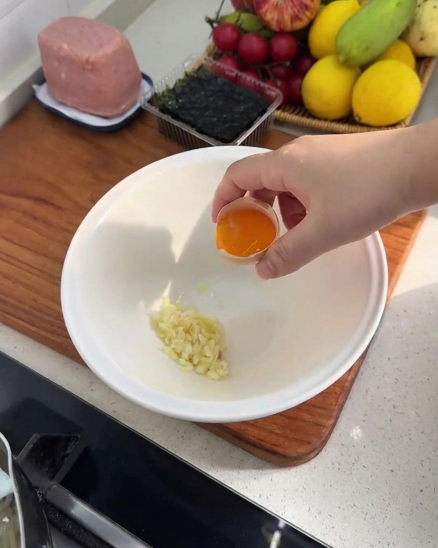 A close-up of a white bowl containing a pile of minced garlic and a single, vibrant egg yolk.