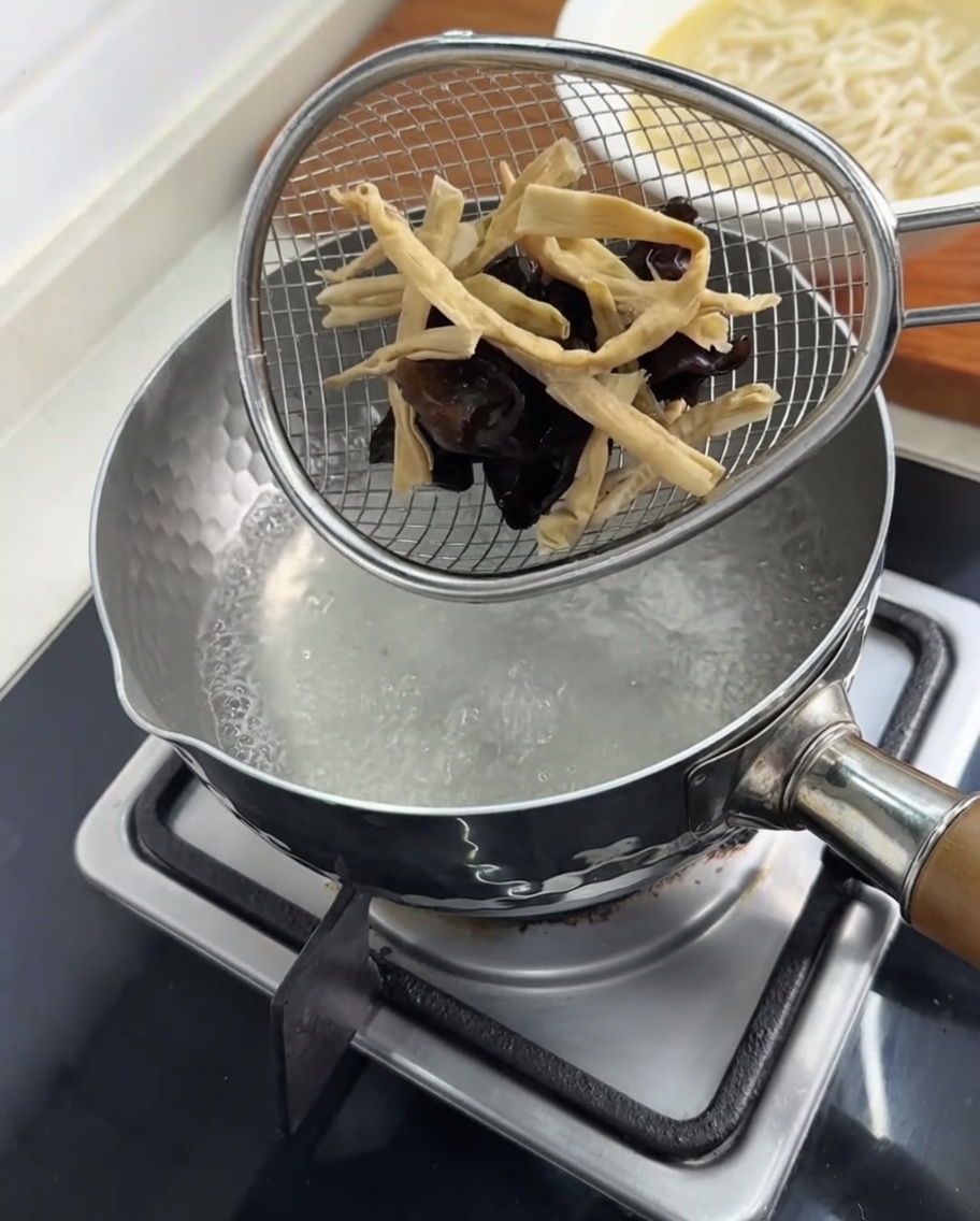 Wood ear mushrooms and bamboo shoots being blanched in a strainer inside a pot of boiling water.