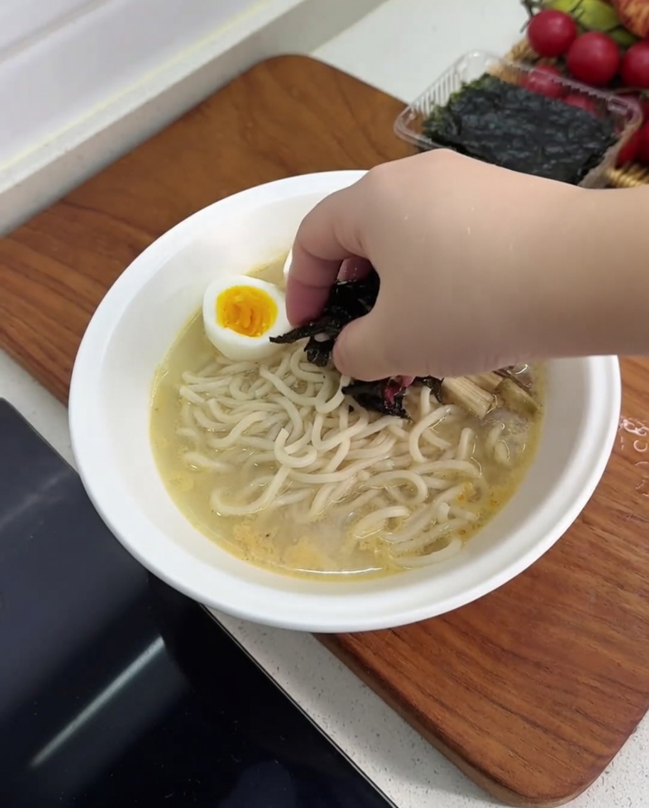 A bowl of ramen being topped with hard-boiled egg halves, mushrooms, bamboo shoots, and a piece of nori seaweed.
