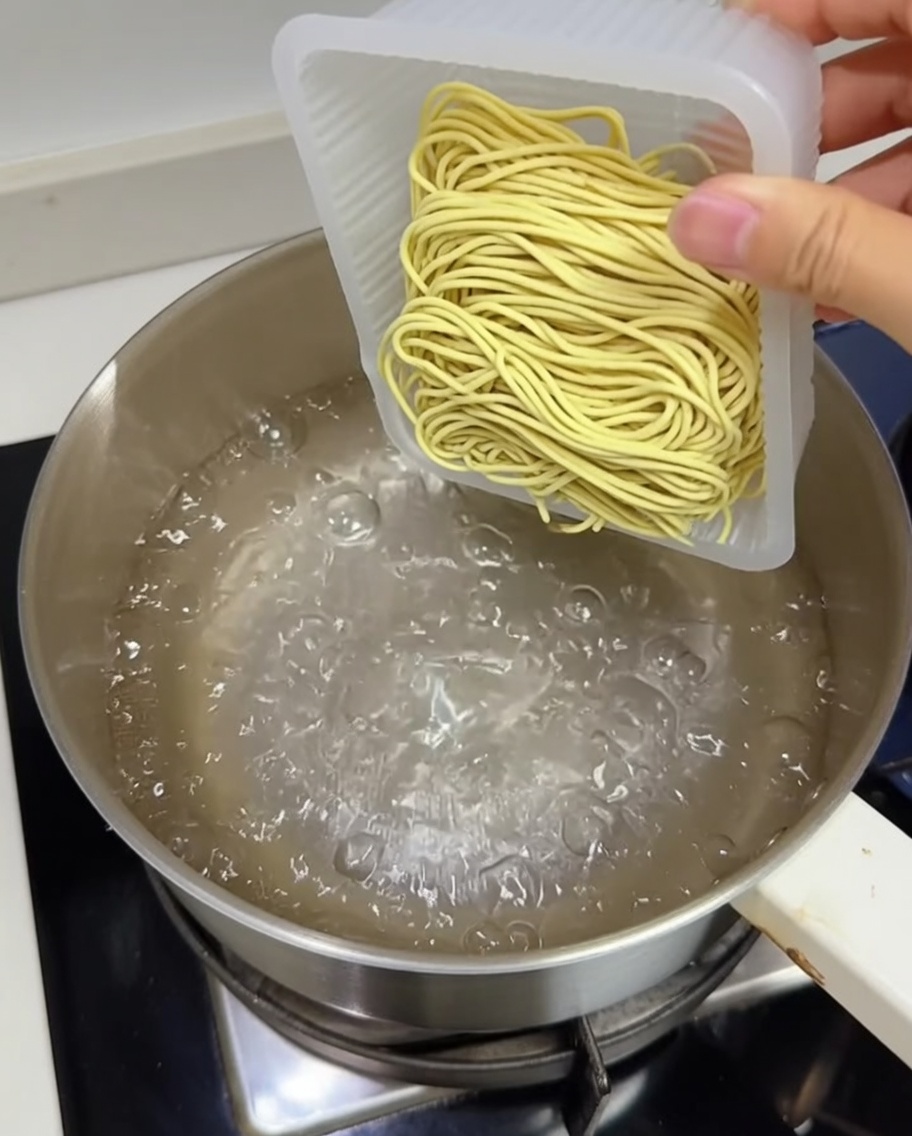 A block of fresh yellow ramen noodles being dropped into a pot of boiling water.
