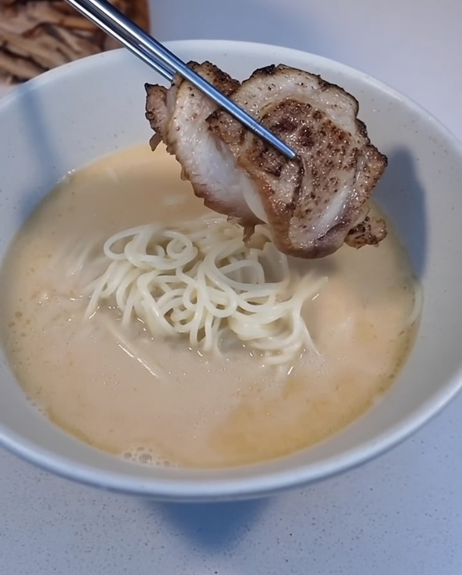 A slice of seared Char Siu being placed onto a bowl of ramen with tweezers.