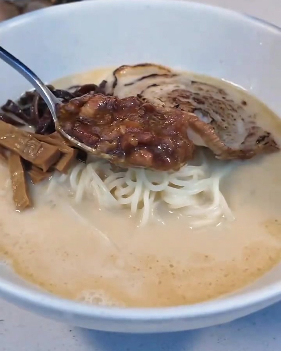 A spoonful of braised meat sauce being placed into the ramen bowl next to the soft-boiled egg half.