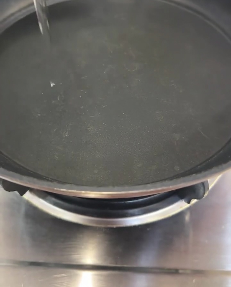 A thin, clear stream of cooking oil being poured into the center of a black frying pan.