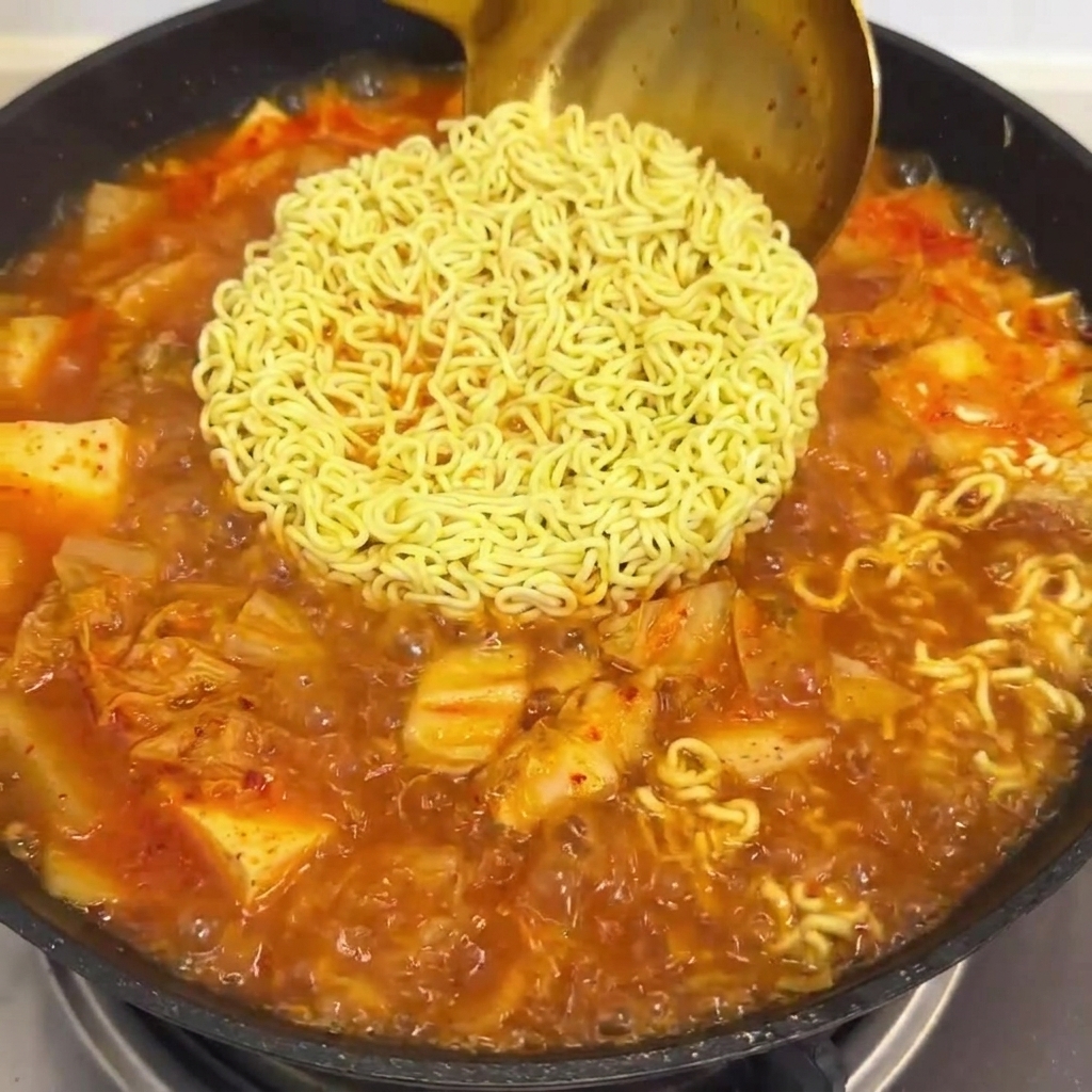 Red sauce being poured into a pan with boiling ramen noodles and kimchi broth.