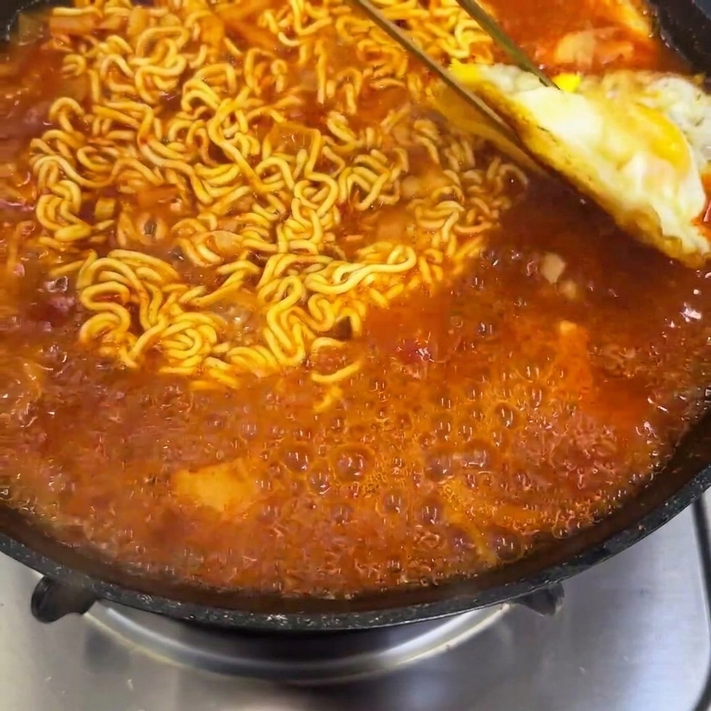 Fried eggs being placed into a pan with simmering spicy ramen noodles.