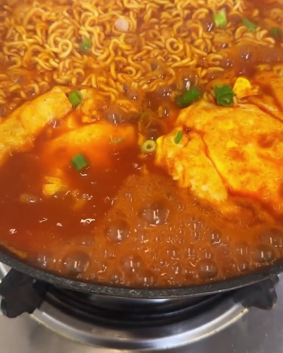 A close-up view of a pot of kimchi ramen topped with freshly scattered green onions and bright red chili slices, with broth bubbling around them.