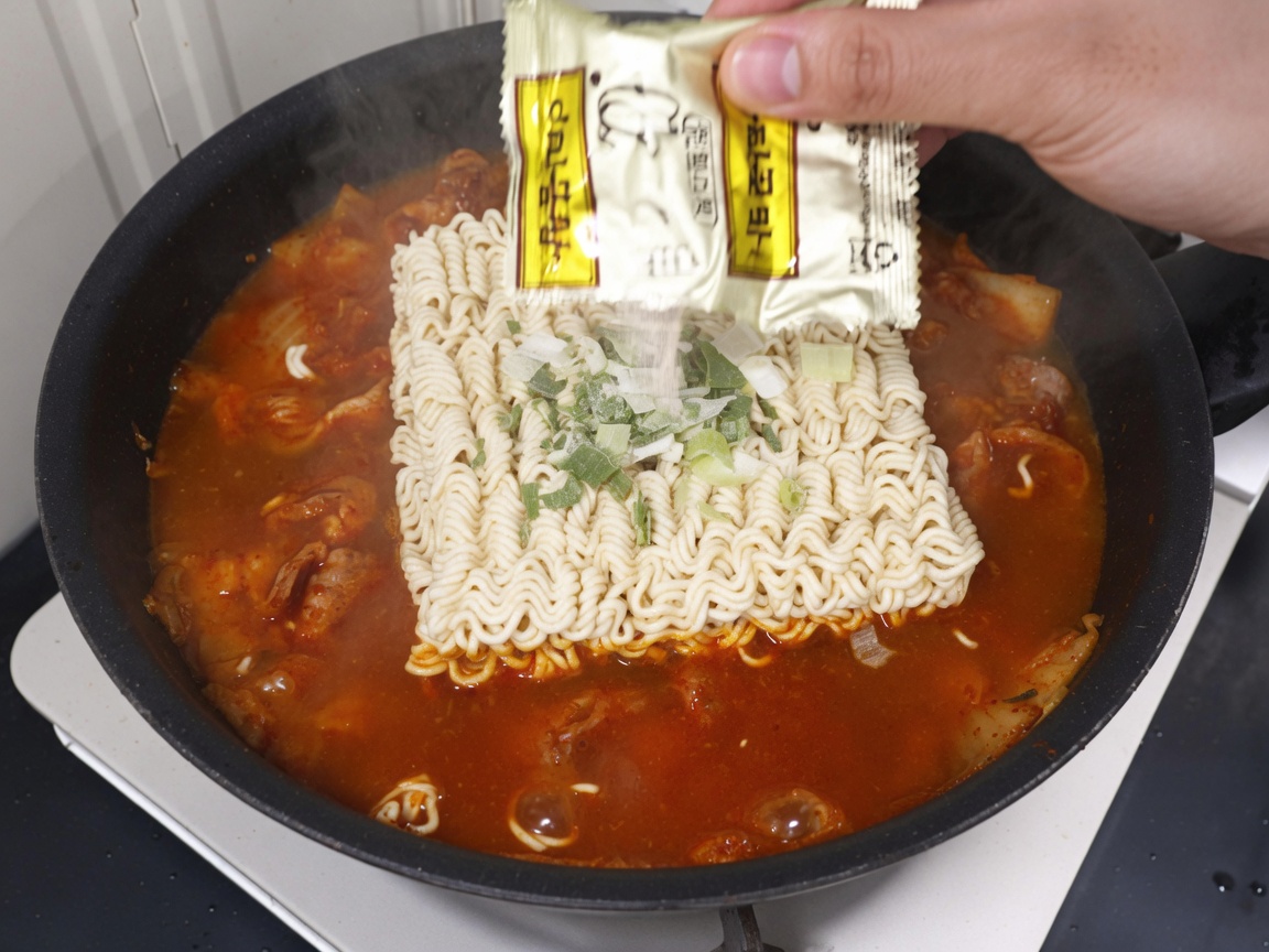 Seasoning packet being poured over the ramen noodles and broth in the pan.