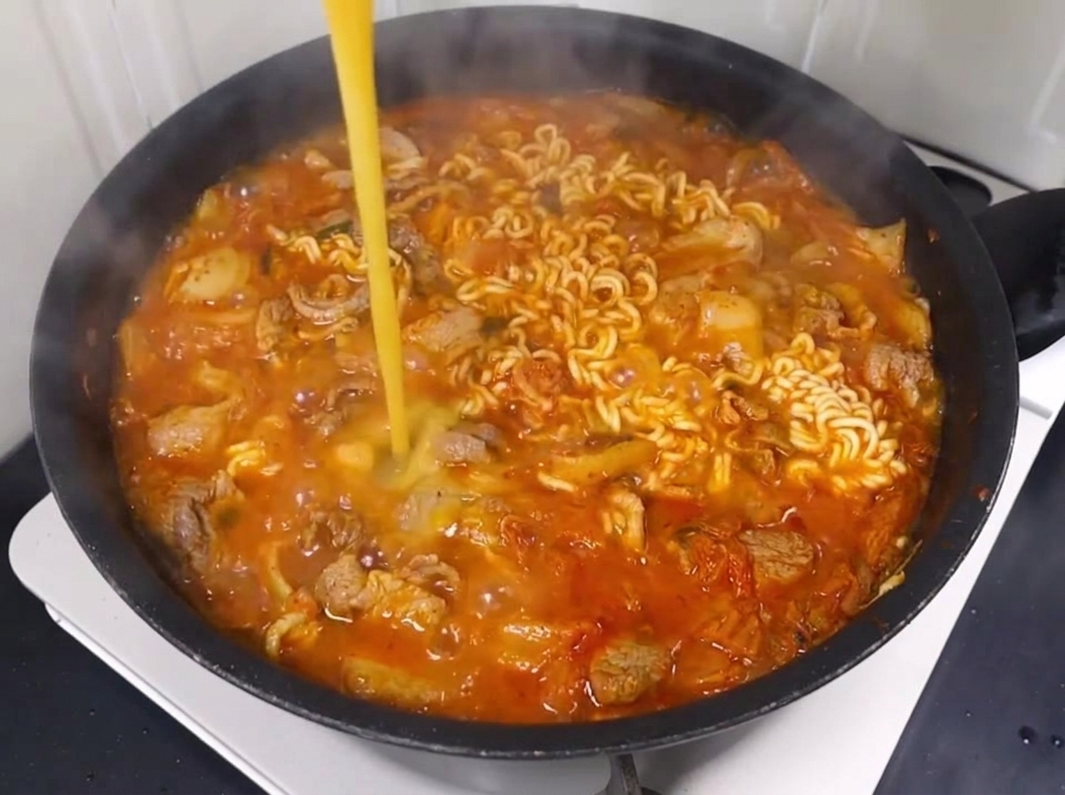 A beaten egg being stirred into the bubbling ramen broth with noodles, pork, and kimchi.