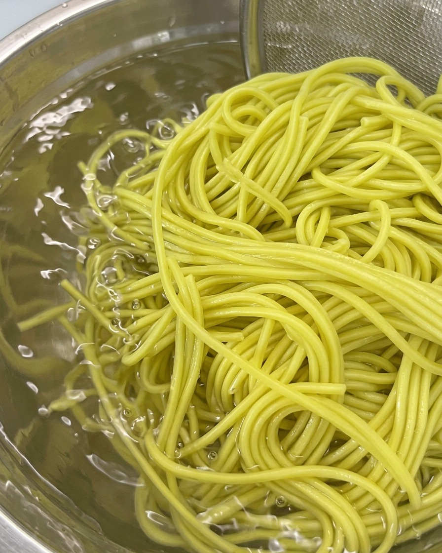 A strainer holding boiled noodles being submerged in a bowl of cold water for rinsing.