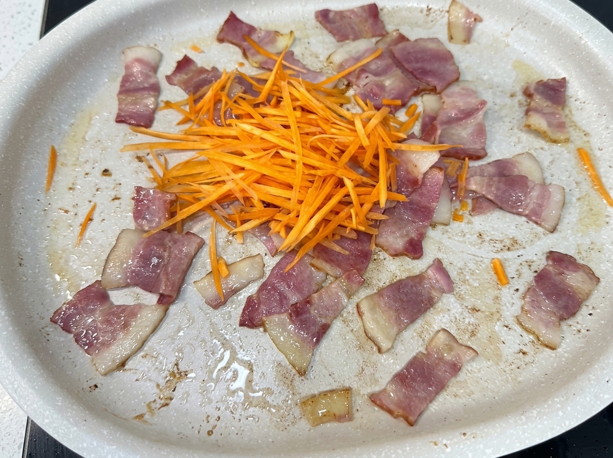 A mix of fresh shredded carrots, onions, cabbage, and bean sprouts being added to a pan with cooked bacon pieces.