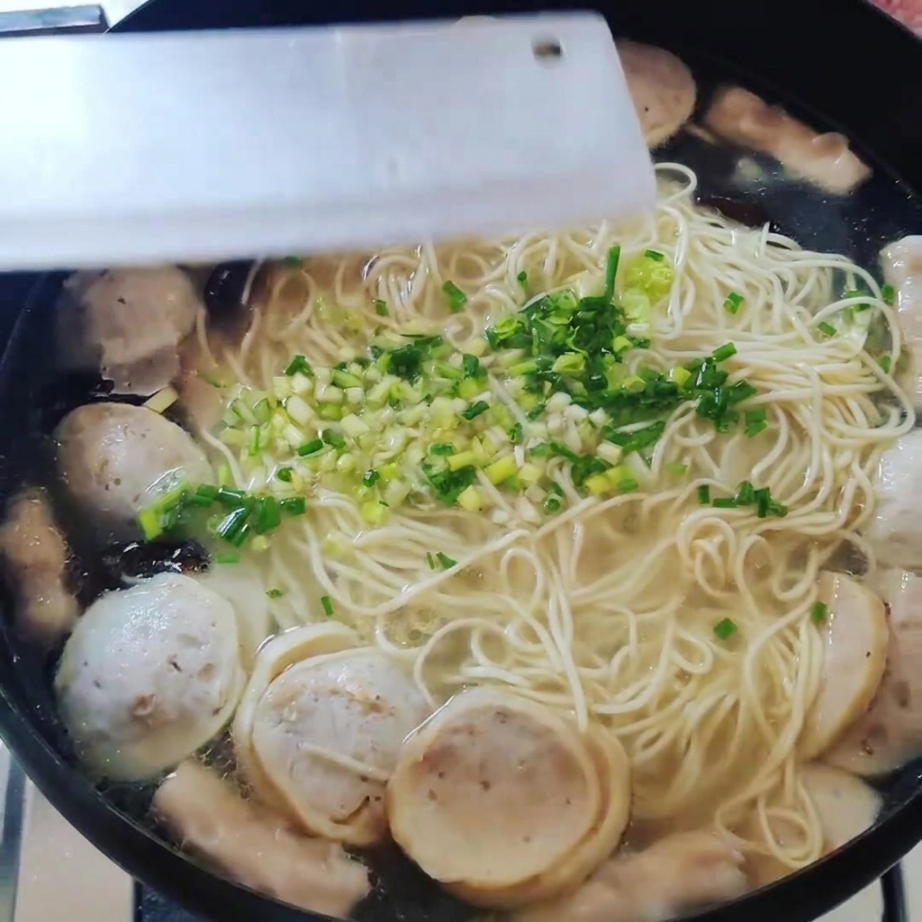 A top-down view of a pot containing noodles, meatballs, and other ingredients being garnished with chopped green onions.