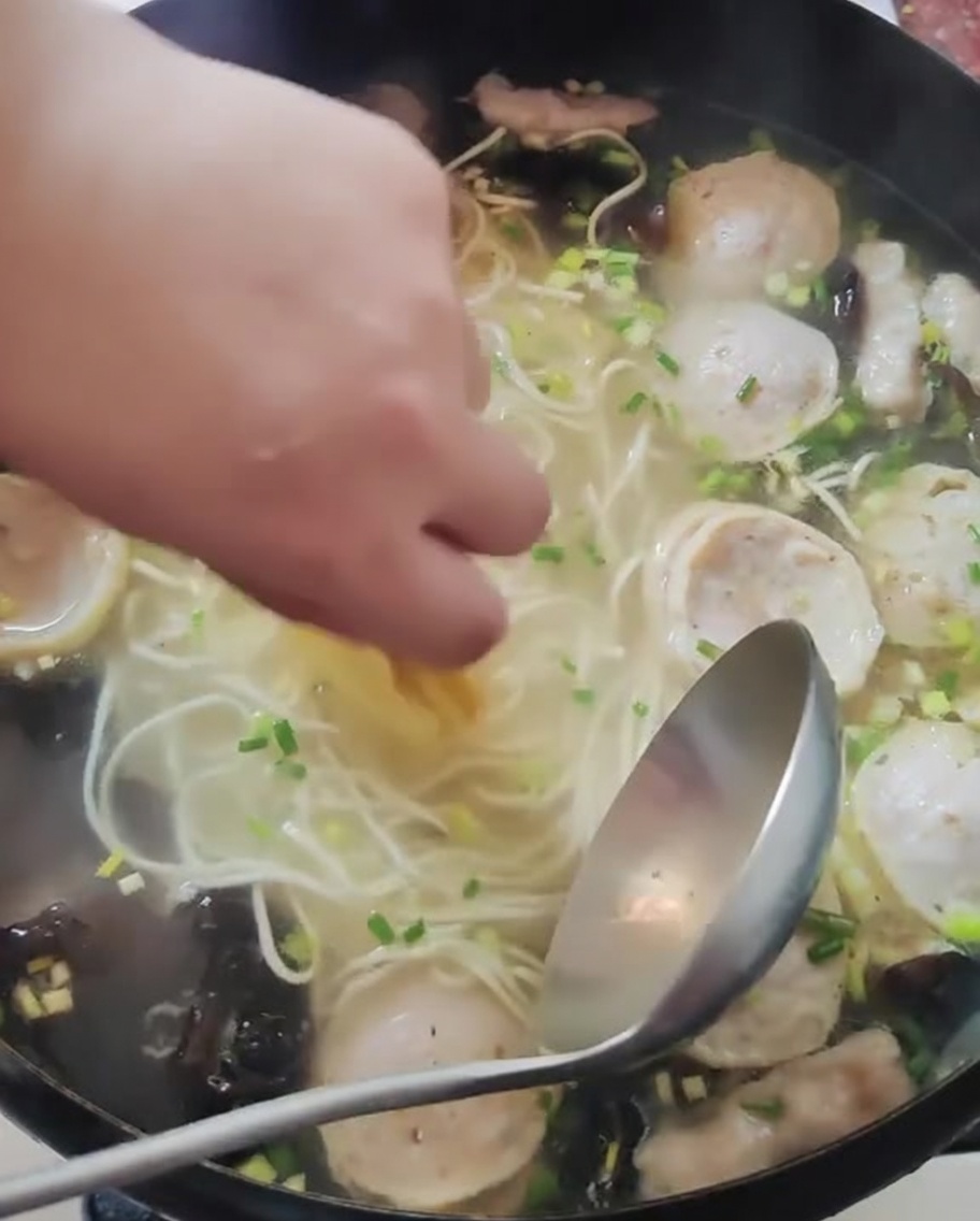 A hand placing thin slices of yellow lemon into a pot of cooked noodles and broth.