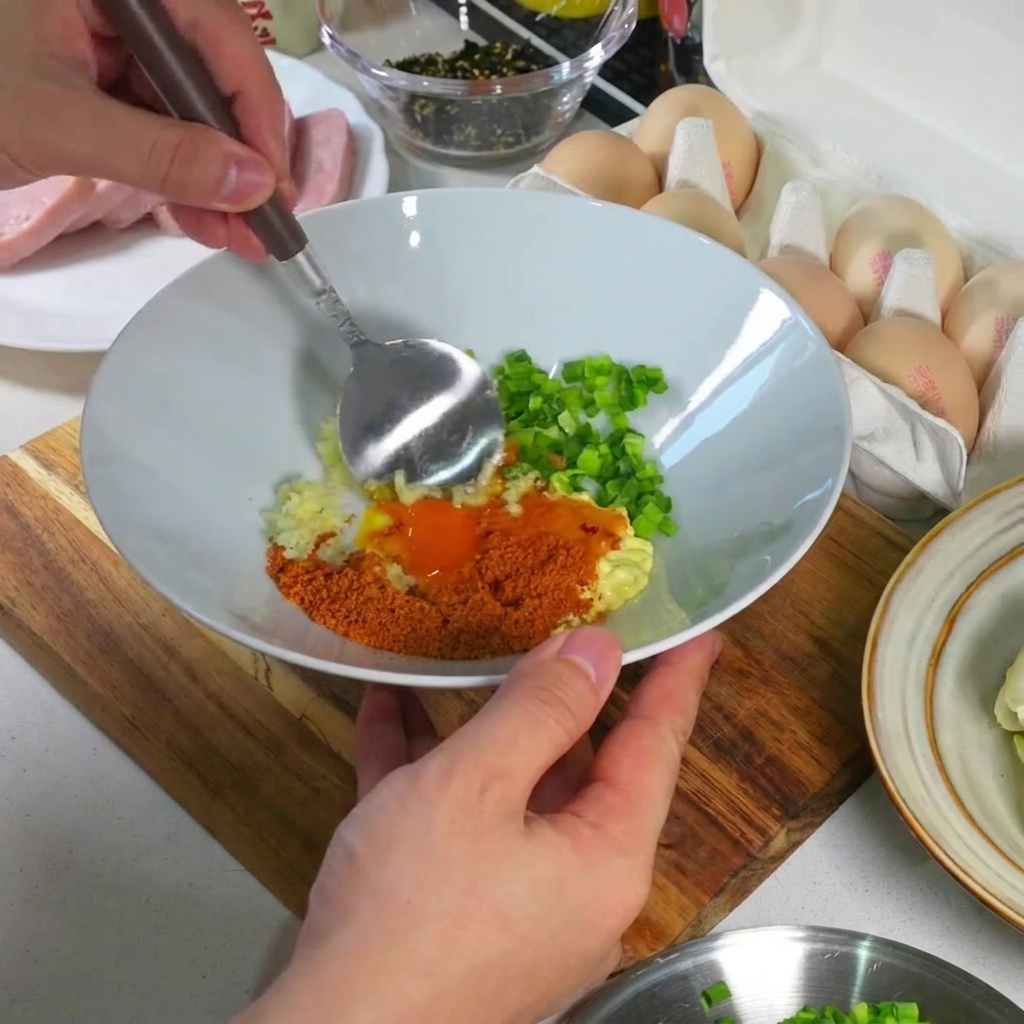 Close-up of a spoon blending the egg yolk, garlic, onions, and seasonings inside a white bowl into a smooth, thick paste.