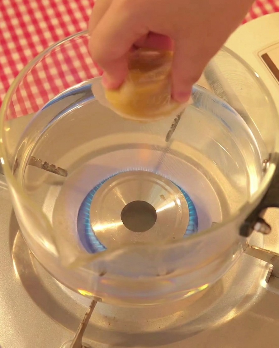 A hand placing a chicken stock cube into a glass pot of boiling water.