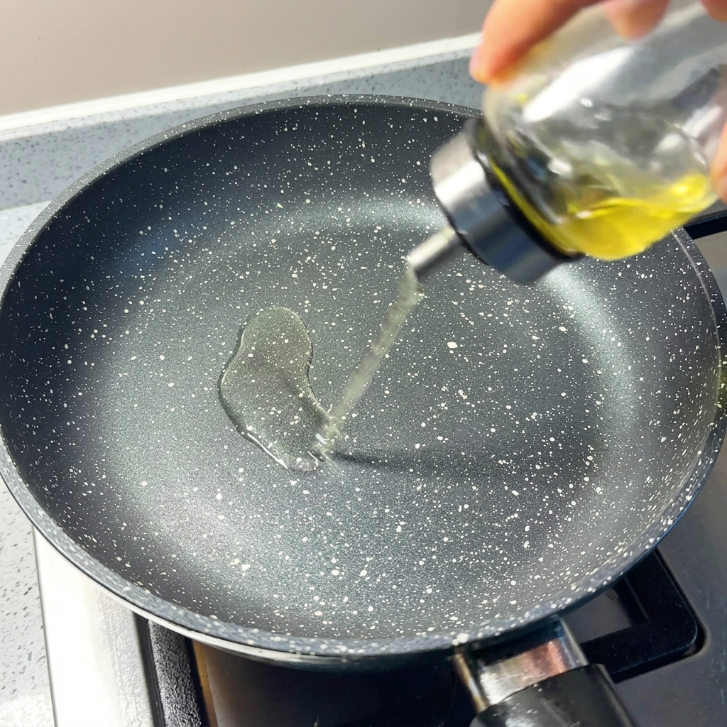 A close-up of clear cooking oil being poured into a preheated, speckled non-stick pan.