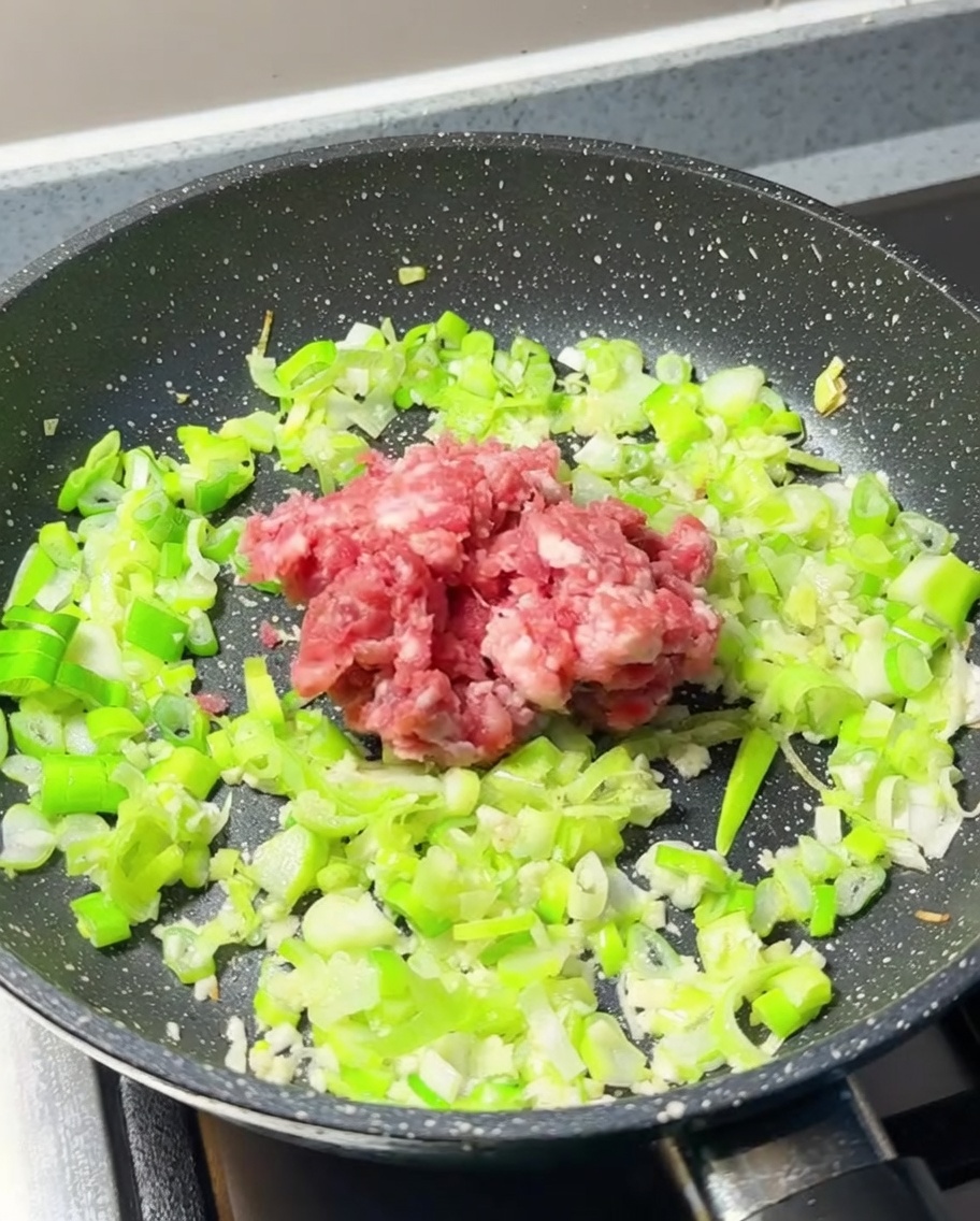 A heap of fresh, raw minced meat added to the center of a pan containing sautéed scallions and garlic.