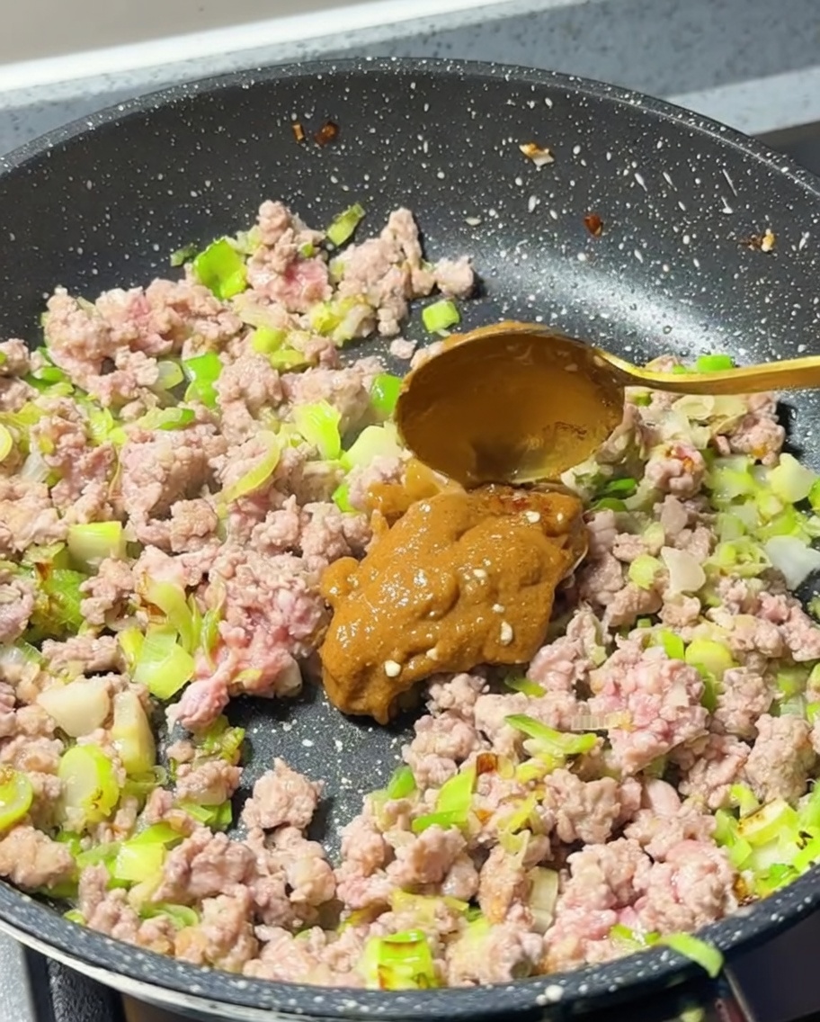 A spoonful of thick, light-brown miso paste being added into the pan with the cooked ground meat.