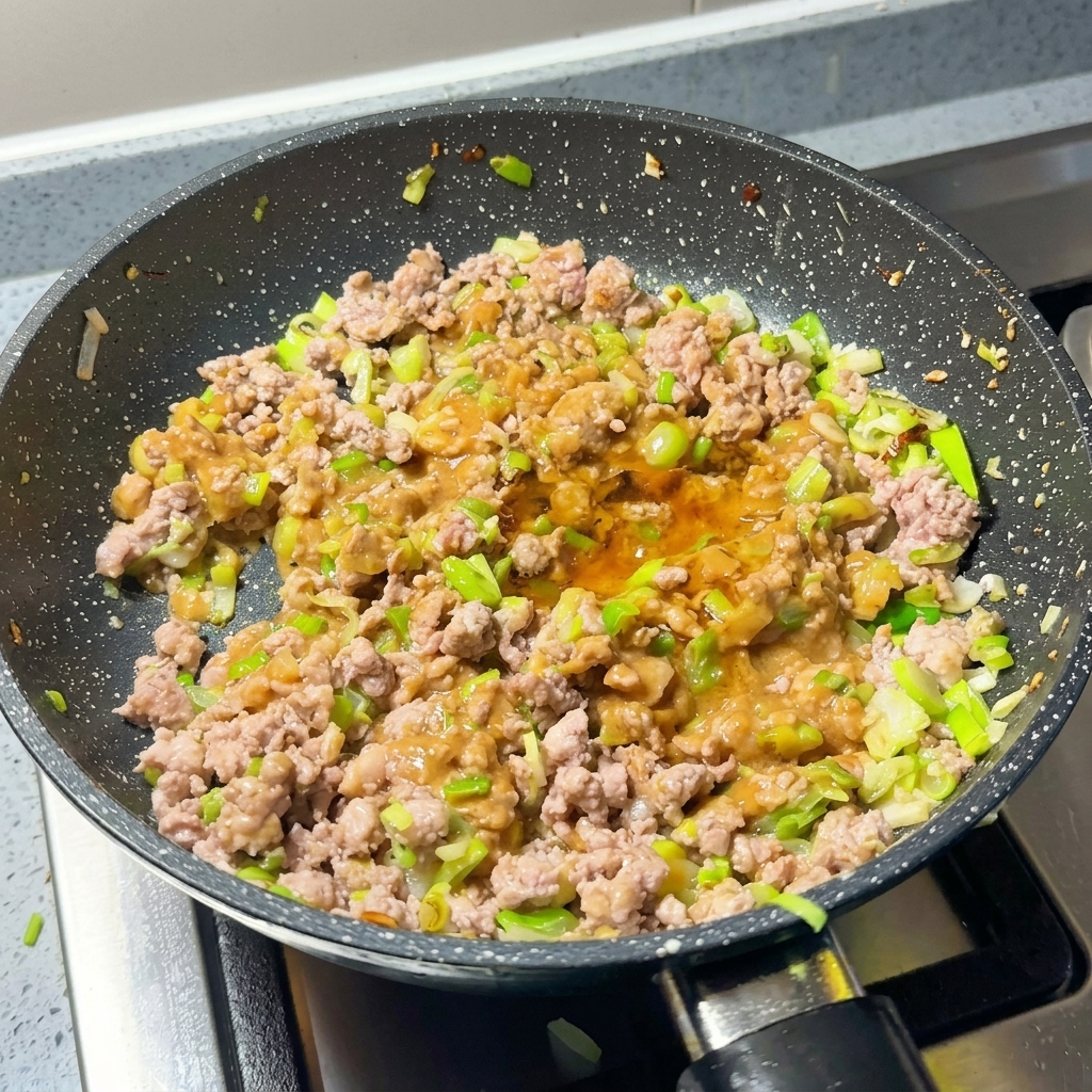 A pan containing browned minced meat, scallions, and miso paste being seasoned with chili powder and sugar.