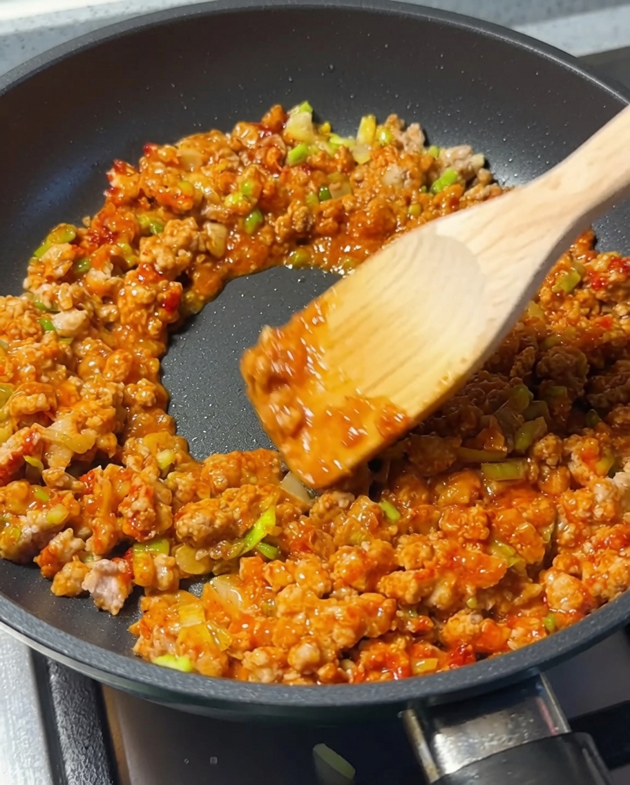 Wooden spoon stirring minced meat and seasonings in a pan as soy sauce is added.