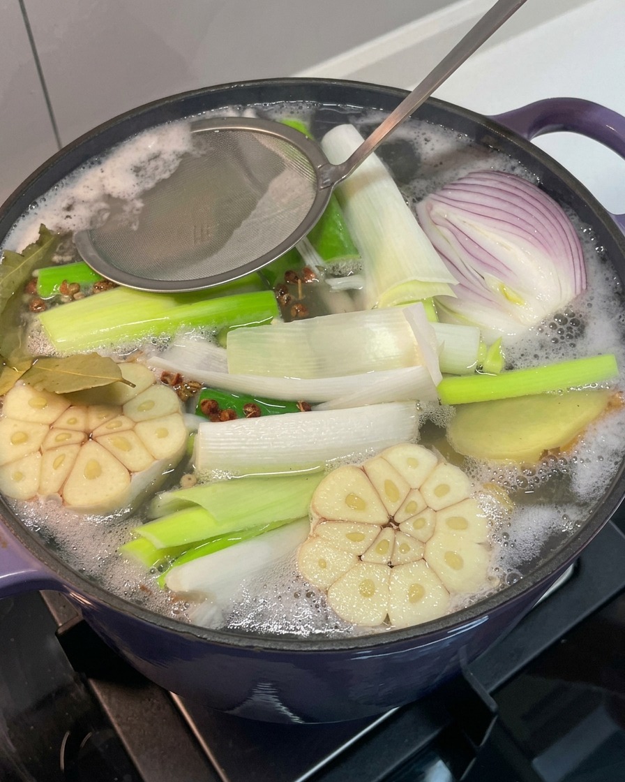 A metal strainer removing gray foam from the surface of the boiling broth.