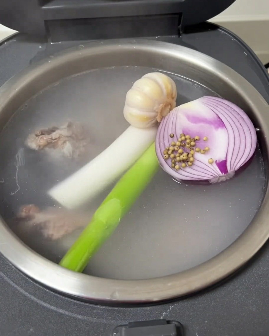 A view inside the rice cooker showing garlic, onion, green onion, and white pepper floating with the pork riblets.