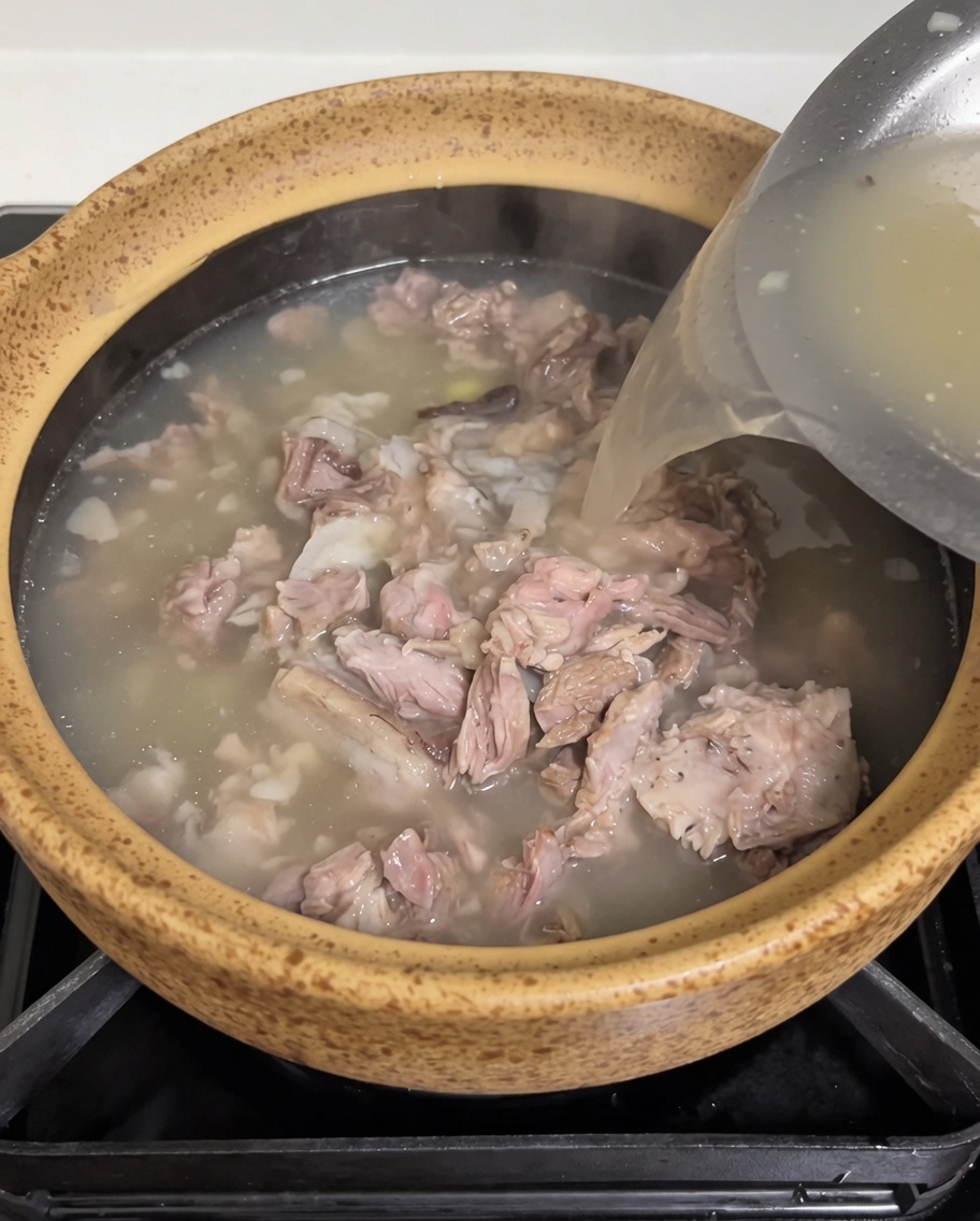 Warm, opaque meat broth being poured from a stainless steel container into the earthen pot containing pork and sprouts.