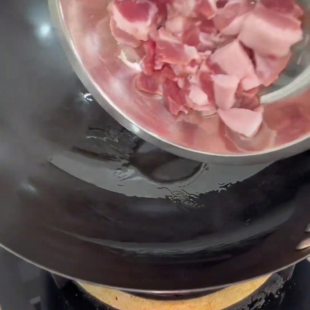 Pork belly slices being added into a hot wok containing a thin layer of cooking oil.