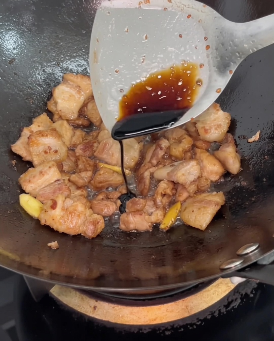 Dark soy sauce being drizzled from a spatula onto the cooking pork belly in the wok to adjust color.