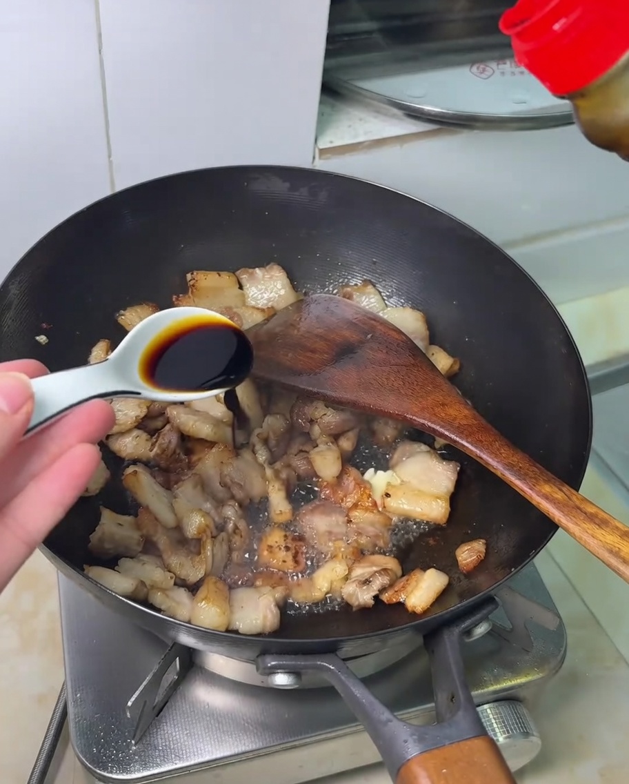 Soy sauce being drizzled over stir-fried pork belly in a wok.