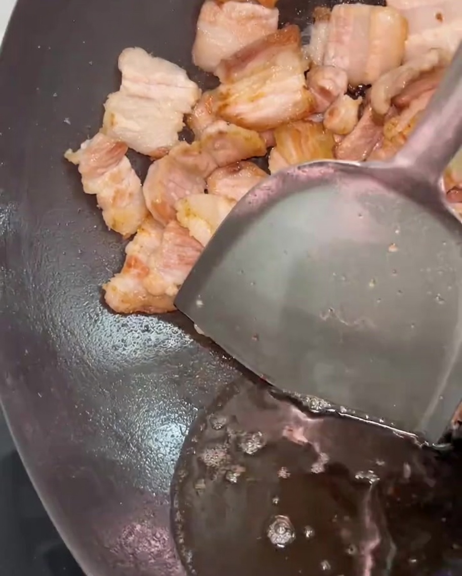 A metal spatula pushing pork belly aside while excess rendered fat is drained from the wok.