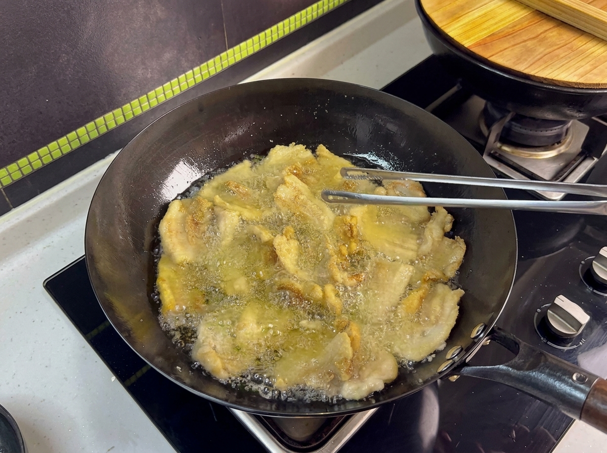 Multiple pork belly slices frying in a wok, being flipped by metal tongs.