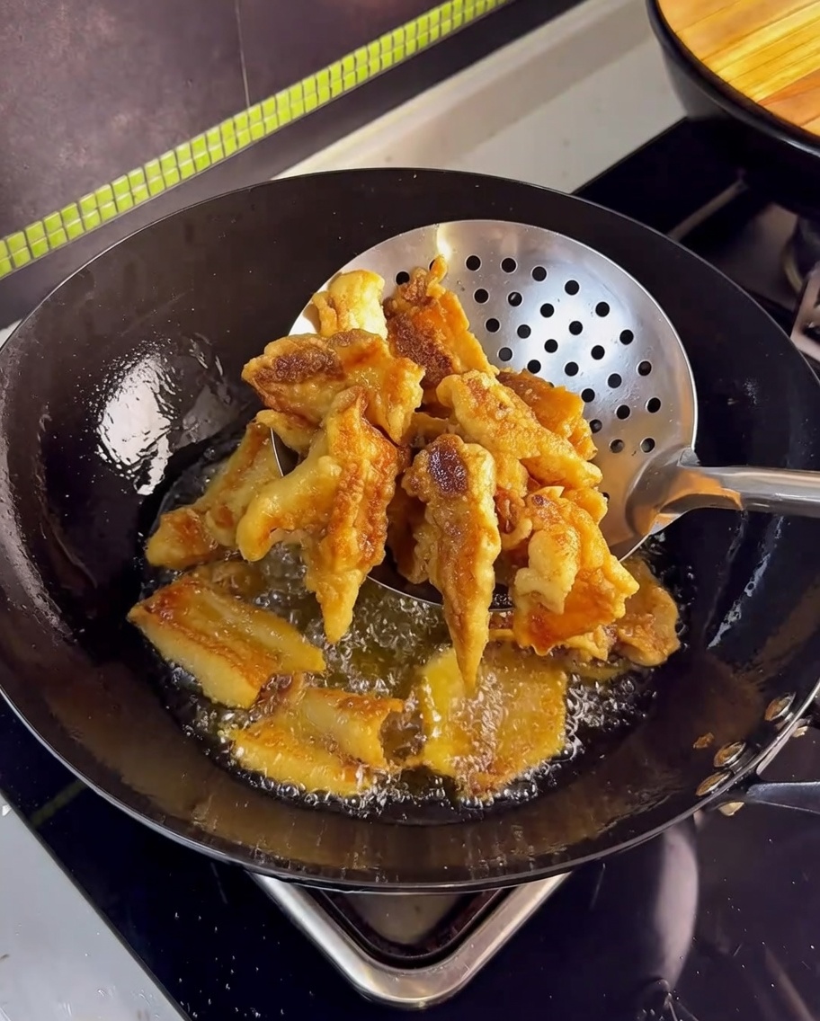 Golden-brown crispy pork belly being lifted out of hot oil with a stainless steel mesh skimmer.