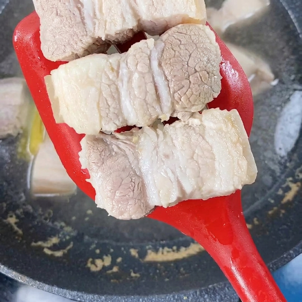 Blanched pork belly pieces being lifted from boiling water with a red spatula.