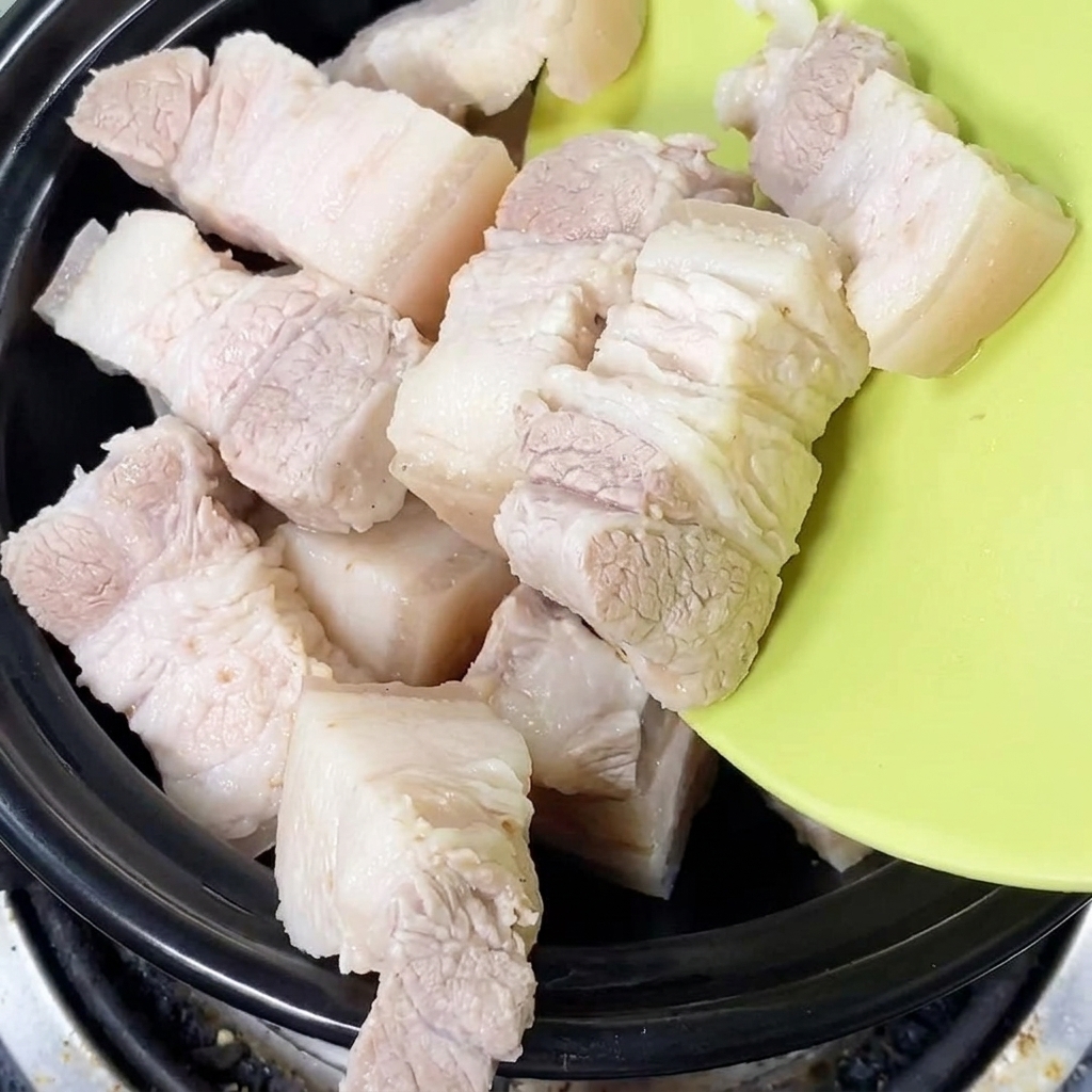 Freshly blanched pork belly pieces being placed into a black clay pot.