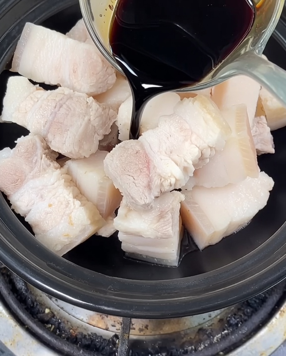 Dark and light soy sauce being poured over pork belly in a clay pot, with rock sugar added.