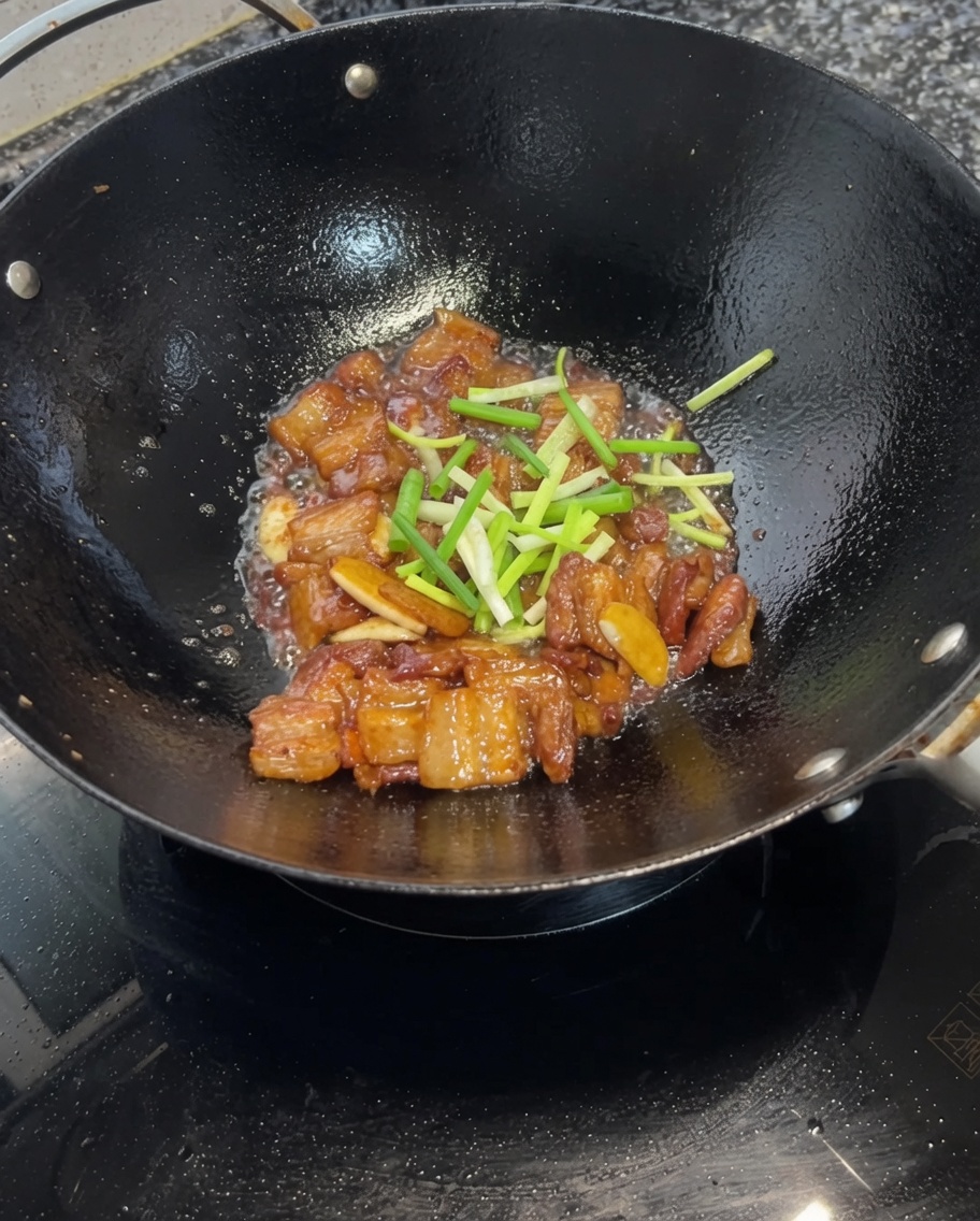 Chopped green onion segments being tossed with the soy-glazed pork belly in the wok.