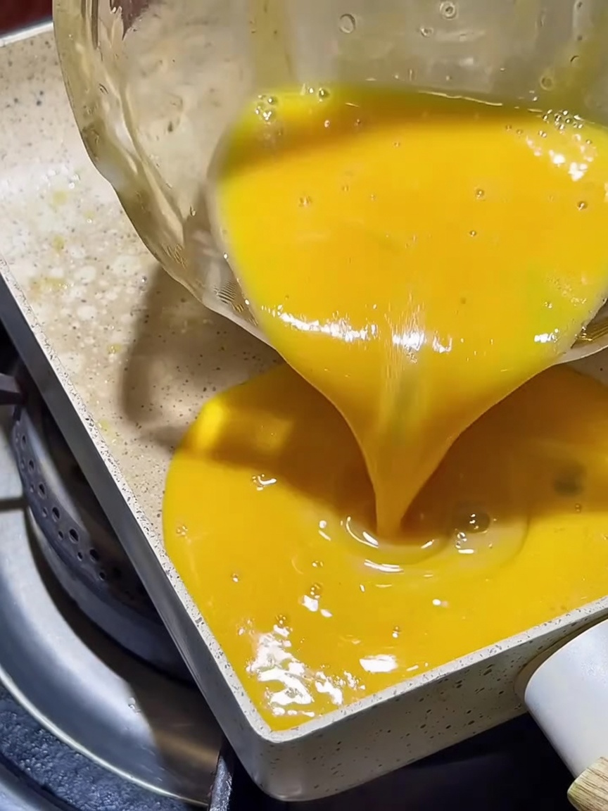 Golden beaten egg mixture spreading evenly in a square non-stick pan.