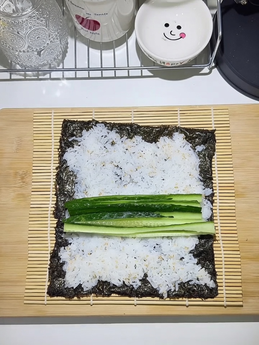 A bamboo mat with a seaweed sheet, spread with rice and layered with cucumber, carrot, egg, and beef strips.
