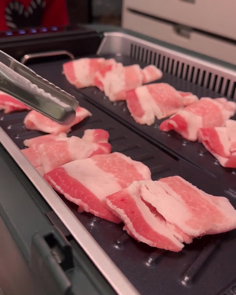 Pork belly slices browning and cooking on an electric grill surface.