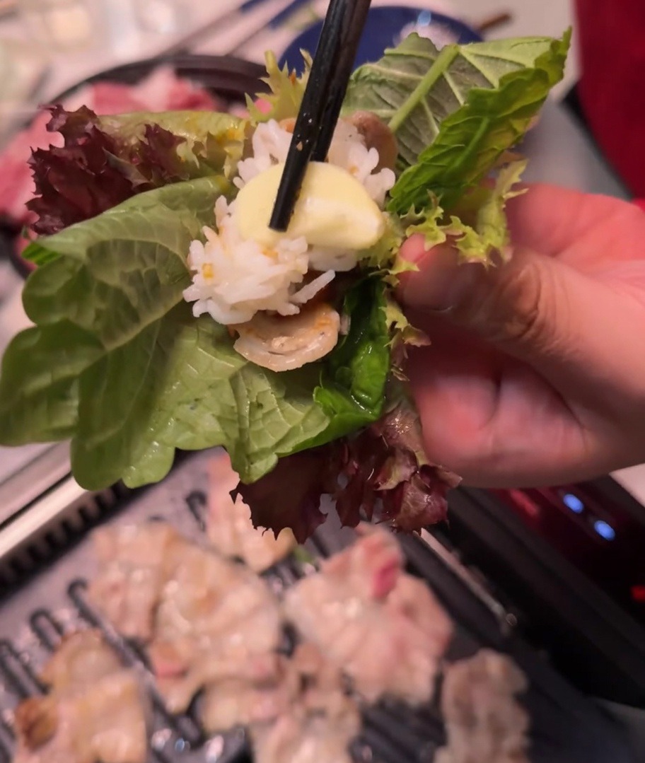 A hand holding a lettuce and perilla leaf wrap containing grilled meat, rice, spicy soybean paste, and a slice of garlic.