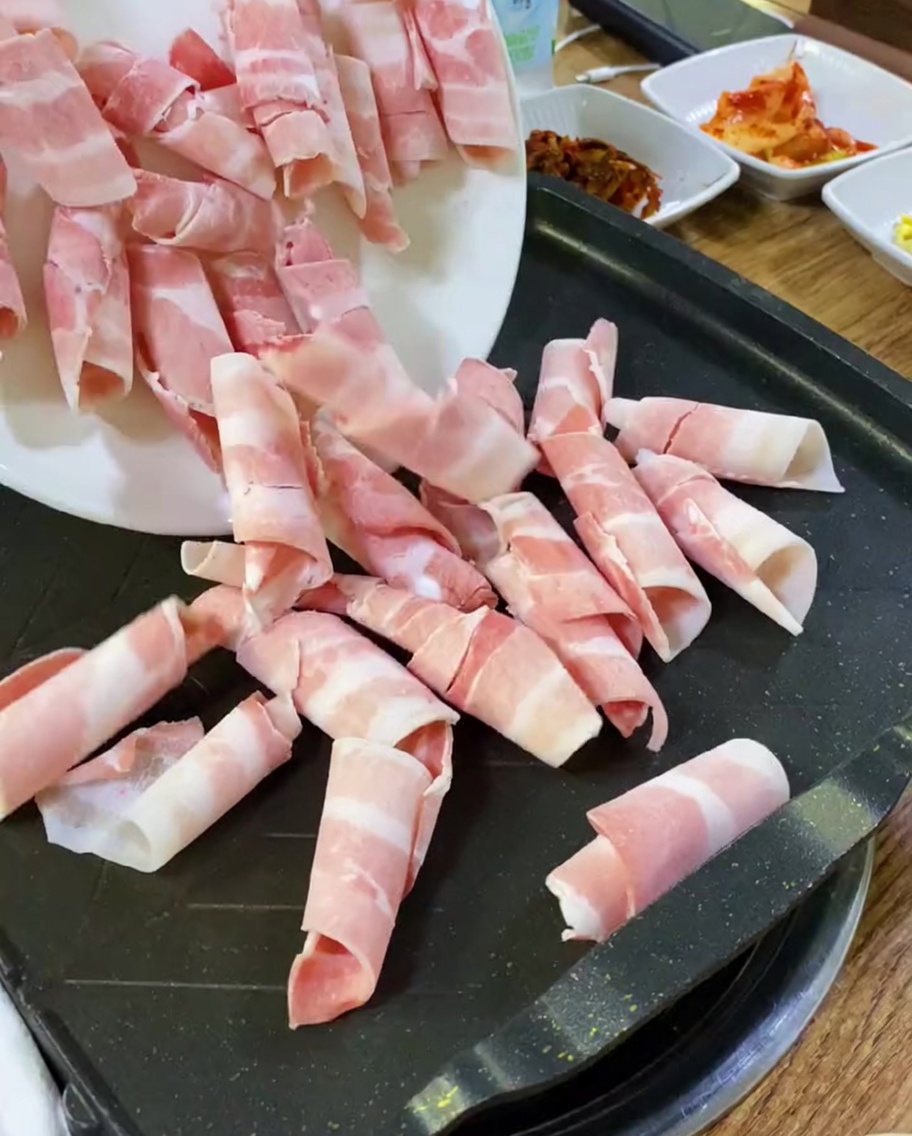 Thin slices of raw pork belly being placed onto a hot griddle.