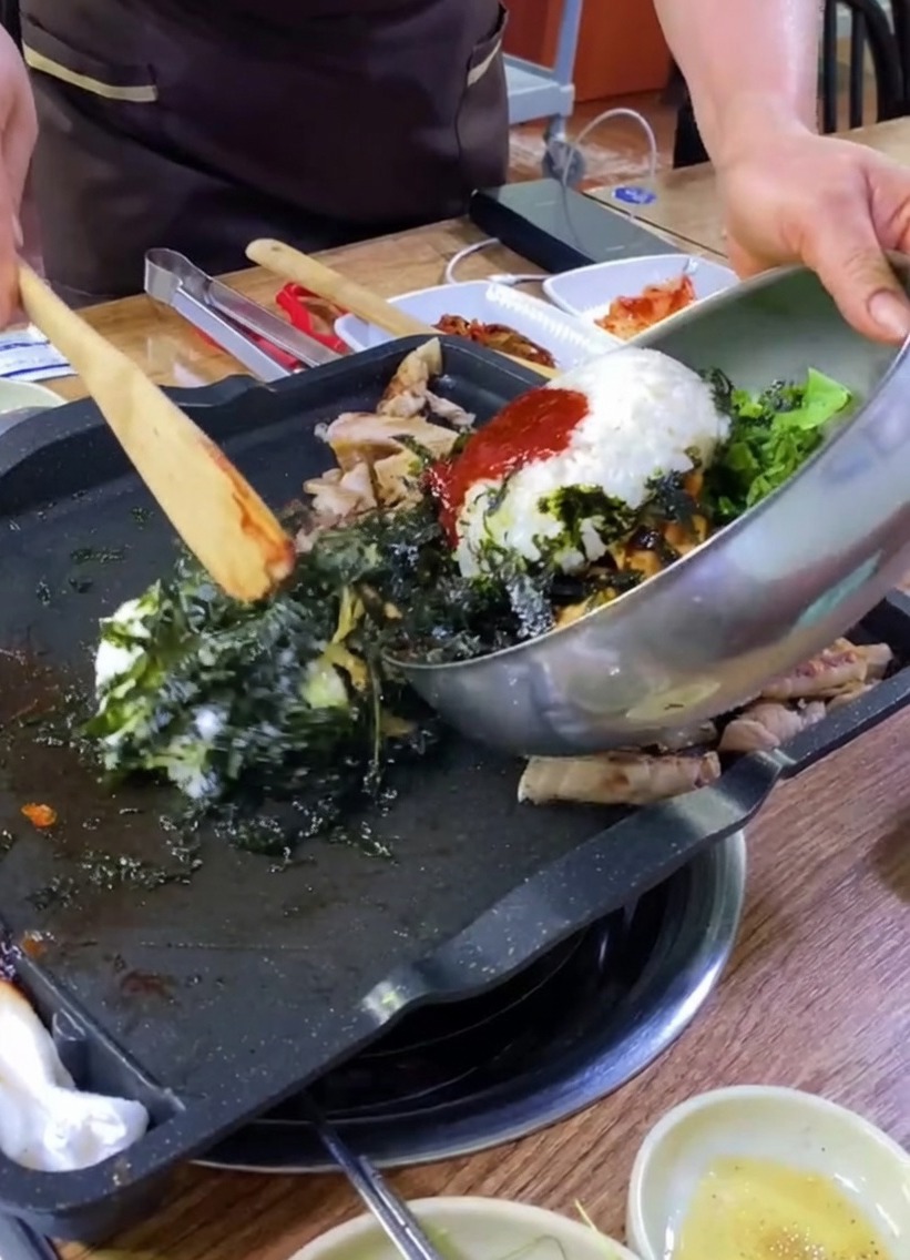 Rice, vegetables, and seaweed being poured from a metal bowl onto a griddle with pork.