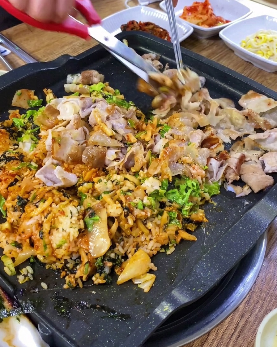 Kitchen shears cutting grilled pork belly into small pieces on a griddle.