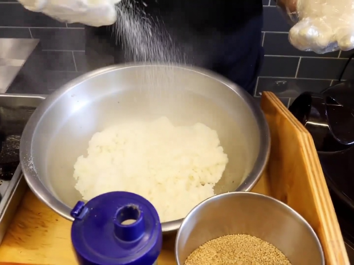 A close-up of a large stainless steel bowl filled with steaming hot sticky rice, with a generous dusting of salt and sesame seeds on top.