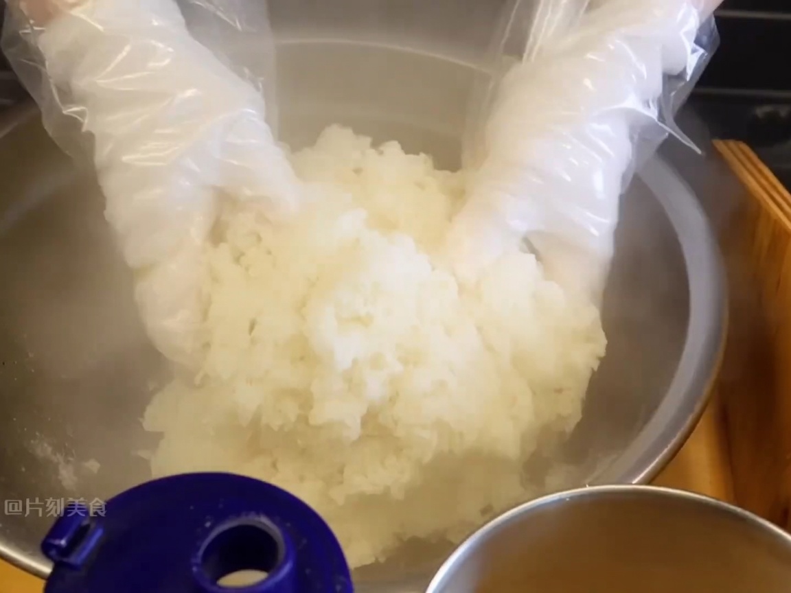 Gloved hands tossing and mixing steaming white sticky rice in a large bowl to incorporate seasonings.