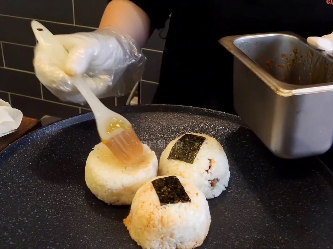 Rice balls sitting on a hot, dark griddle being brushed with a rich, brown glaze by a gloved hand.